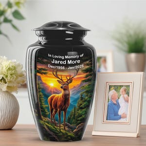 May include: A black cremation urn with a glossy finish, featuring a scenic sunset and deer illustration. The text "In Loving Memory of Jared More Dec/1956 - Jan/2025" is displayed. A framed photo is next to the urn.