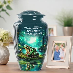 Peut inclure: Urne funéraire turquoise avec une illustration d'une grenouille dans un étang, avec une pleine lune en arrière-plan. L'urne porte l'inscription "In Loving Memory of Jared More Dec/1956 - Jan/2025". Une photo encadrée est à côté.
