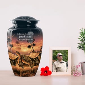 May include: A black cremation urn featuring a sunset beach scene with palm trees and a sea turtle. The text reads In Loving Memory of Jared More Dec/1966 - Jan/2026. The urn has a smooth finish and a matching lid, displaying a warm orange glow.