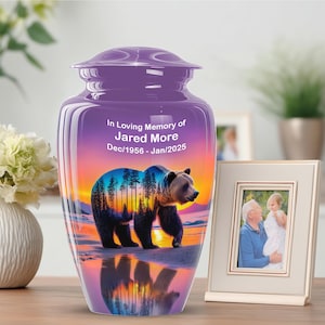 May include: A purple cremation urn with a bear and sunset scene. The urn has the text "In Loving Memory of Jared More Dec/1956 - Jan/2025". A framed photo is next to the urn.