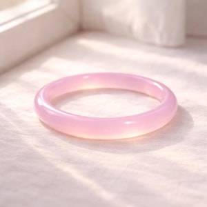 May include: A light pink, circular bangle bracelet. The smooth, polished surface reflects light, creating a soft glow. The bracelet is made of a translucent material, and is set against a white background.