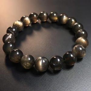 Op de afbeelding: Een cirkelvormige armband bestaande uit gladde, ronde, donkergrijze kralen. Elke kraal heeft een reflecterende, chatoyante glans die een glinsterend, metaalachtig effect op het oppervlak van de steen creëert, gerangschikt in een doorlopende lus.