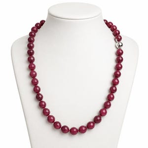 Op de afbeelding: Een ketting bestaande uit ronde, gefacetteerde, diep magenta gekleurde kralen die aan elkaar zijn geregen. De ketting is voorzien van een enkele, gladde, gepolijste zilverkleurige bolvormige sluiting aan de rechterkant die de kralenreeks vastzet.