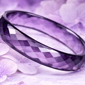 May include: A purple bangle bracelet with a geometric, faceted design. The bracelet is a solid band of amethyst-coloured material. The background features soft purple flowers, complementing the bracelet's colour and design.