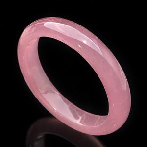 Puede incluir: Un brazalete rosa claro y pulido. El brazalete redondeado está hecho de un material translúcido, que refleja la luz. La joya está sobre un fondo negro. El diámetro del brazalete es de aproximadamente 6,5 cm.