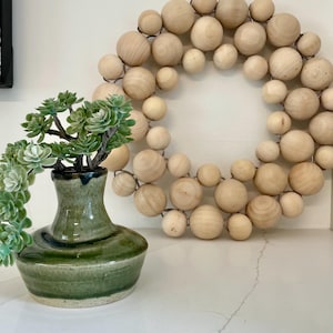 May include: A green ceramic vase with a succulent plant and a wooden bead wreath. The vase has a dark green and blue glaze. The wreath is made of light-coloured wooden beads strung together with wire. Home decor.