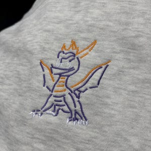 Puede incluir: Sudadera gris claro con un dise&ntilde;o de drag&oacute;n bordado. El drag&oacute;n est&aacute; delineado con hilo morado y naranja, con detalles blancos. El drag&oacute;n est&aacute; representado con las alas extendidas, mirando hacia la derecha.