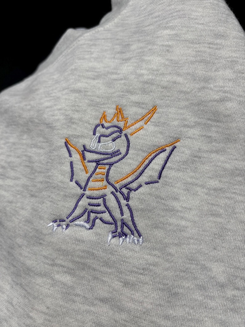 Puede incluir: Sudadera gris claro con un dise&ntilde;o de drag&oacute;n bordado. El drag&oacute;n est&aacute; delineado con hilo morado y naranja, con detalles blancos. El drag&oacute;n est&aacute; representado con las alas extendidas, mirando hacia la derecha.