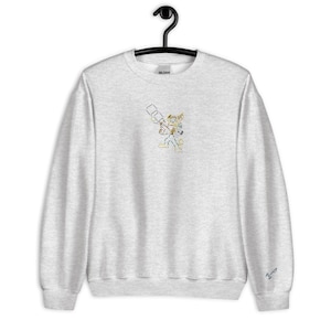 May include: A light gray sweatshirt with a crew neck and long sleeves. The front features a colorful embroidered design of a cartoon character holding a weapon. The sweatshirt is hanging on a black hanger.
