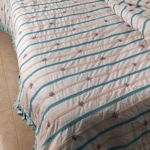 May include: A white bedspread featuring teal and beige horizontal stripes, embellished with small embroidered starbursts. The bedspread has a teal ruffled edge, creating a coordinated aesthetic. This bedding set is designed for a double or queen size bed.