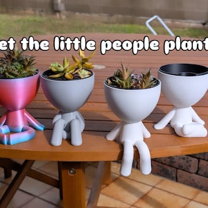 May include: Four quirky planters in the shape of little people, each containing a succulent. The planters are in shades of red, blue, and white. The text "Meet the little people planters" is displayed above the planters.