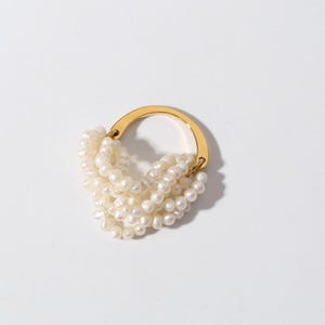 Puede incluir: Un anillo dorado con múltiples hilos de perlas blancas. El anillo tiene un diseño circular, con las perlas cayendo en cascada desde la parte superior. Las perlas están agrupadas, creando un efecto de capas. El anillo está sobre un fondo blanco.