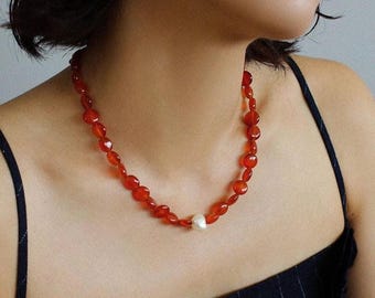 May include: A necklace composed of small, polished, translucent orange-red beads arranged in a circular strand. A single, irregular, off-white pearl is positioned at the center of the necklace, serving as a focal point among the vibrant red beads.
