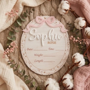 Peut inclure: Un panneau de naissance ovale en bois avec un nœud 3D rose clair sur le dessus. Le texte indique Sophie ROSE avec des lignes vides pour Date, Time, Weight et Length. Le panneau est orné d'une bordure délicate de fleurs et de papillons.
