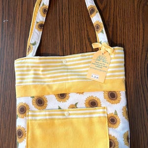 May include: A tote bag with a sunflower design. The bag has a white background with yellow sunflowers and green leaves. It features a yellow pocket and yellow and white striped accents. The handles are white with sunflower patterns.