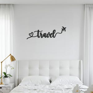 May include: Black metal wall art spelling "Travel" with a heart and airplane design. The decorative piece is mounted on a white wall above a white bed with pillows. A gold lamp and white flowers are on a nightstand.