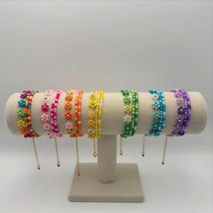May include: A display of seven beaded bracelets in various colors, each featuring a floral design. The bracelets are arranged on a white cylindrical stand, showcasing a spectrum of hues including pink, orange, yellow, green, blue, and purple.