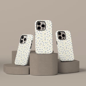 May include: Three phone cases with a floral pattern. The cases are white with a repeating pattern of small blue flowers and green leaves. The cases are displayed on a neutral-colored, geometric display.