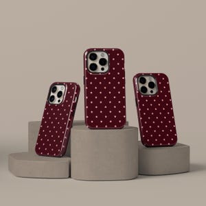 May include: Three maroon phone cases with a repeating pattern of small, light pink stars. The cases are displayed on a neutral-toned, geometric platform. The cases vary in size, suggesting different phone models.