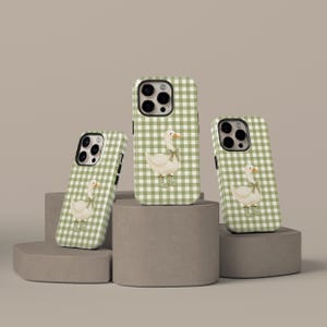 Puede incluir: Tres fundas para teléfono con un patrón de cuadros verdes y blancos. Cada funda presenta un ganso blanco con un lazo verde. Las fundas se exhiben sobre pedestales geométricos grises sobre un fondo neutro.