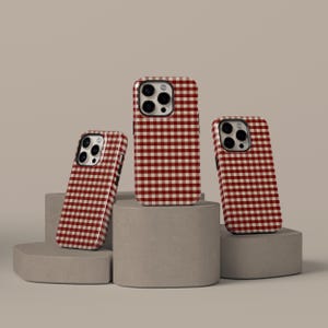 Peut inclure: Trois coques de téléphone avec un motif vichy rouge et blanc. Les coques sont présentées sur des socles géométriques gris sur un fond neutre. Les coques ont des découpes pour l'appareil photo et une finition brillante.