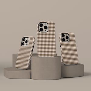Può includere: Tre custodie per telefono con un motivo a quadretti marrone e bianco sono esposte su piedistalli geometrici grigi. Le custodie sono progettate per adattarsi a uno smartphone, con ritagli per la fotocamera e i pulsanti. Le custodie sono di colore neutro.