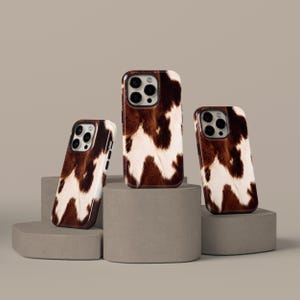 May include: Three mobile phone cases featuring a brown and white cowhide design. The cases are presented on grey cylindrical plinths against a neutral backdrop. The cases have a glossy finish and a dark brown border.