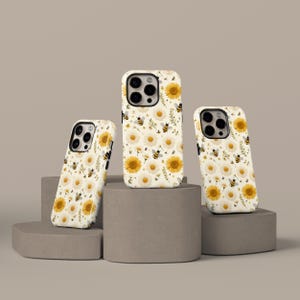 May include: Three phone cases with a floral design featuring white daisies, yellow sunflowers, and buzzing bees. The cases are on a neutral background, showcasing the detailed pattern and design. The cases are in different sizes.