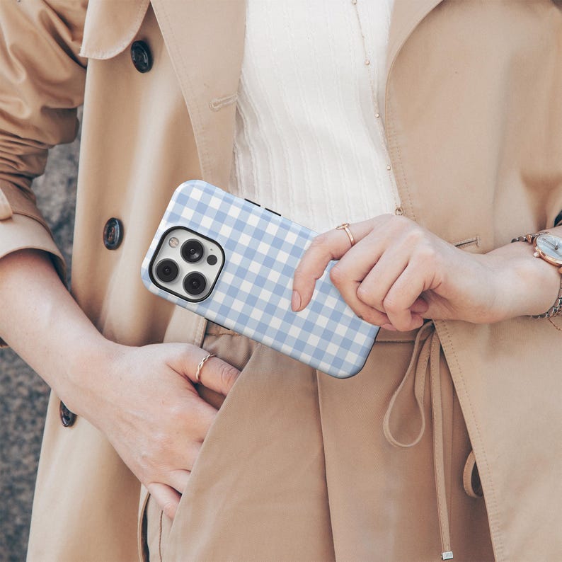 May include: A light blue and white gingham patterned phone case. The phone case is held in a person's hand, and the case has a black border. The phone case is a modern design.