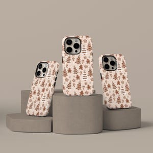 May include: Three phone cases with a festive Christmas pattern. The cases feature a repeating design of gingerbread men, Christmas trees, snowflakes, and candy canes on a light beige background. The cases are displayed on gray geometric blocks.