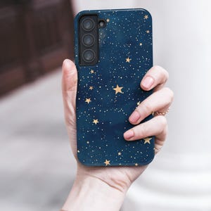 May include: A navy blue smartphone case featuring a celestial design with gold stars and white dots. The case has a triple-lens camera cutout. The phone case is held in a hand, with a rose gold watch visible.