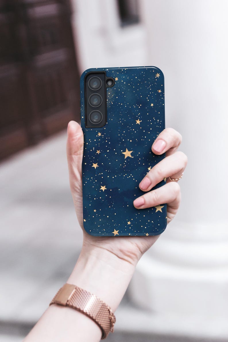 May include: A navy blue smartphone case featuring a celestial design with gold stars and white dots. The case has a triple-lens camera cutout. The phone case is held in a hand, with a rose gold watch visible.