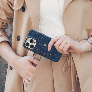 May include: A navy blue phone case with a gold star pattern is held in front of a tan trench coat. The phone case has a camera cutout and a black border. The person holding the phone case is wearing rings and a watch.