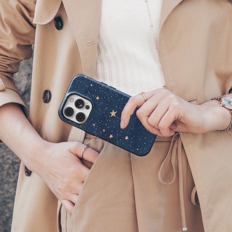 May include: A navy blue phone case with a gold star pattern is held in front of a tan trench coat. The phone case has a camera cutout and a black border. The person holding the phone case is wearing rings and a watch.