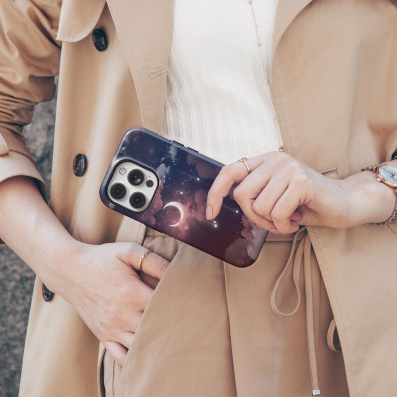 Puede incluir: Funda de tel&eacute;fono azul oscuro con un dise&ntilde;o celestial que presenta una luna creciente, estrellas y nubes. La funda se sostiene en una mano, con un abrigo largo beige en el fondo.
