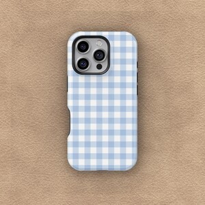 May include: A light blue and white gingham pattern phone case. The case has a camera cutout and button cutouts. The phone case is on a tan surface.