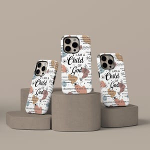 May include: Three phone cases with a white background, featuring the text "I am a Child of God" in a script font. The cases have floral and abstract shapes in muted colors. The cases are on a neutral-toned surface.