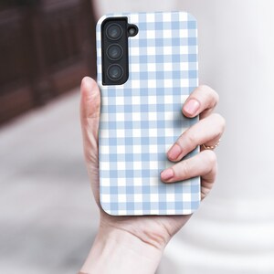 May include: A light blue and white gingham patterned phone case. The case has a rectangular shape with rounded corners and a camera cutout. The phone case is held in a person's hand.