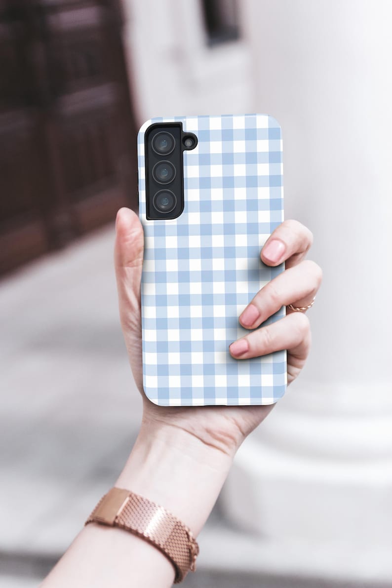 May include: A light blue and white gingham patterned phone case. The case has a rectangular shape with rounded corners and a camera cutout. The phone case is held in a person's hand.