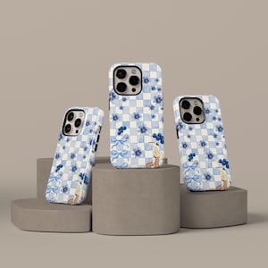 Pode incluir: Três capas de telemóvel com um padrão xadrez azul e branco, adornadas com flores azuis, laços e ilustrações. As capas são exibidas em plataformas geométricas cinzentas sobre um fundo neutro.