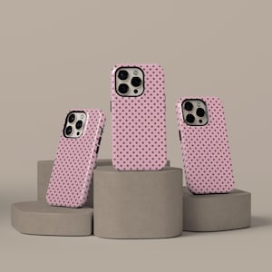 May include: Three pink phone cases with a repeating pattern of dark pink polka dots. The cases are displayed on gray geometric pedestals against a neutral background. The cases have camera cutouts and button placements.