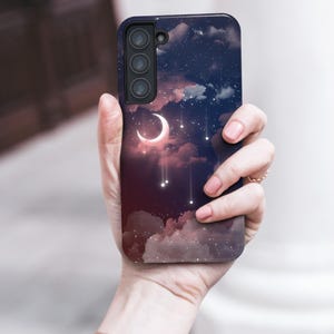 Puede incluir: Funda de tel&eacute;fono con dise&ntilde;o celestial, con una luna creciente, estrellas y nubes sobre un fondo azul oscuro y rojo. La funda tiene un m&oacute;dulo de c&aacute;mara negro y se sostiene en una mano.