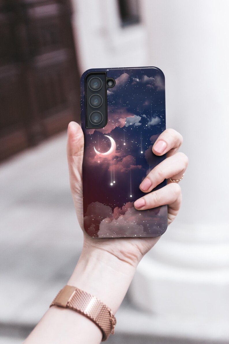 Puede incluir: Funda de tel&eacute;fono con dise&ntilde;o celestial, con una luna creciente, estrellas y nubes sobre un fondo azul oscuro y rojo. La funda tiene un m&oacute;dulo de c&aacute;mara negro y se sostiene en una mano.