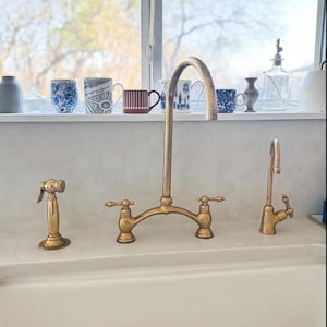 Könnte beinhalten: Ein Messing-Küchenarmaturen-Set mit einem hohen, gebogenen Auslauf, zwei Hebelgriffen, einer seitlichen Brause und einem separaten, kleineren Wasserspender. Die Armaturen haben eine metallische Oberfläche und stehen auf einer weißen Arbeitsplatte.
