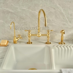 Puede incluir: Un juego de grifos de cocina de color dorado con diseño de puente, que incluye un grifo de filtro de agua separado y un pulverizador lateral. El juego está instalado sobre un fregadero doble blanco. El grifo tiene un estilo clásico y elegante.