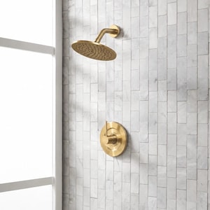 May include: A brushed gold shower system featuring a circular shower head with a perforated face and a matching single-handle mixing valve control. The hardware is mounted on a wall covered in vertical rectangular white marble tiles with gray veining.