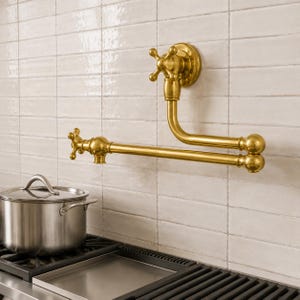 May include: A wall-mounted pot filler faucet with a polished brass finish, featuring two articulated arms and two cross-style handles. The faucet is positioned above a stainless steel cooking pot on a black stovetop with a tiled backsplash.