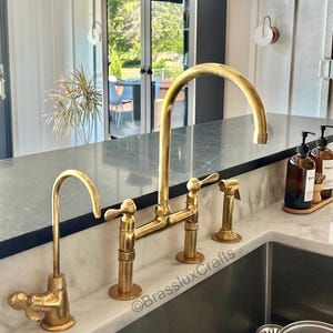 May include: A gold-coloured kitchen tap set with a high-arc spout, side sprayer, and separate water filter tap. The tap is installed on a marble countertop with a stainless steel sink.
