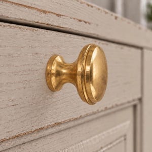 May include: A polished brass cabinet knob with a rounded, mushroom-shaped head and a tapered base, mounted onto a light gray, distressed wooden drawer front with a visible wood grain texture and worn edges showing a darker brown finish underneath.