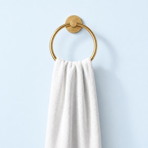 May include: A circular gold-toned metal towel ring mounted to a wall, featuring a smooth, matte finish. A clean, white, plush fabric towel is draped through the center of the ring, hanging vertically with soft, natural folds in the fabric.